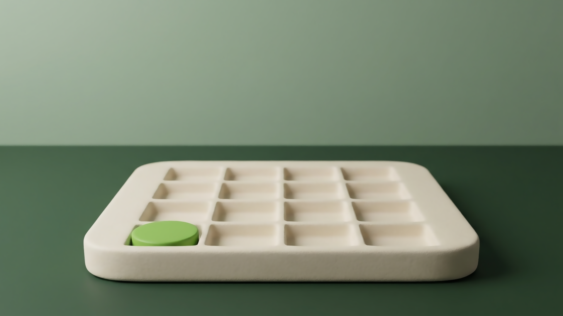 A cream tray on a green desk divided into a grid of small empty square wells, with one green disc resting in a single well at the front edge of the grid